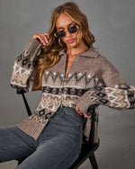 Woman in a brown Fair Isle zip-up cardigan with collar, gold sunglasses and gray high-waisted jeans, seated on a black chair
