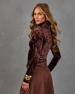 Woman in a brown crushed velvet turtleneck top and wide-leg brown trousers with chunky gold bangles, looking over her shoulder