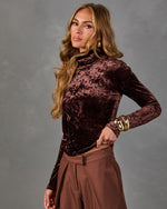 Woman in a brown crushed velvet turtleneck top and wide-leg brown trousers with chunky gold bracelets and earrings