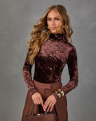 Woman in a brown crushed velvet turtleneck top and brown trousers with gold bangles, holding a brown suede handbag