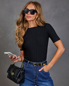 A woman wearing a black short-sleeve ribbed knit top, blue jeans, black sunglasses, and carrying a black quilted handbag, posed against a textured gray studio background.