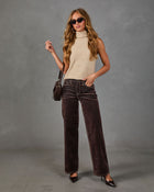 A woman modeling a sleeveless beige knit mock neck top, dark brown wide-leg velvet pants, black pointed-toe heels, and carrying a small black shoulder bag against a textured gray studio backdrop.