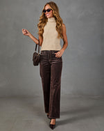 A woman modeling a sleeveless beige knit turtleneck sweater, dark brown wide-leg velvet pants, and black pointed-toe heels, accessorized with a small dark brown handbag and sunglasses, against a textured gray studio background.