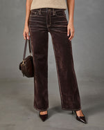A model wearing dark brown wide-leg velvet pants, styled with a beige top, dark brown pointed-toe heels, and carrying a dark brown textured shoulder bag against a gray studio background.