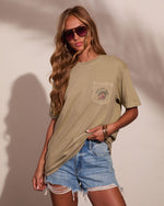 A woman modeling an oversized khaki green pocket t-shirt with a graphic on the chest, paired with distressed denim shorts, set against a warm beige wall with soft leaf shadows.