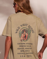 A woman shown from the back wearing a beige graphic T-shirt with a vintage 'Wild West Rodeo' design featuring a horse illustration and multiple destination cities listed below.