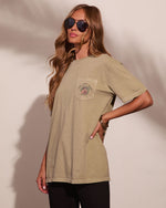 A woman modeling a relaxed-fit, short-sleeve pocket t-shirt in a sandy beige color. The shirt features a chest pocket with a circular graphic that reads 'SUNRISE' and 'SUNSET'.