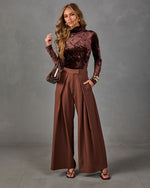 Woman in brown crushed velvet turtleneck and wide-leg pleated chocolate trousers with strappy heels and a brown mini handbag