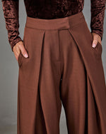 Brown wide-leg pleated trousers with asymmetric wrap overlay and high waistband, styled with a crushed velvet long-sleeve top