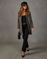 Woman in herringbone tweed coat, black bodysuit, flare jeans, baker boy cap, and pointed heels carrying a black woven handbag