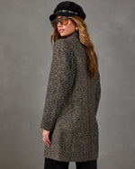 Woman in a brown herringbone tweed coat, black newsboy cap and amber aviator sunglasses, seen from behind against gray wall
