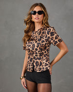 A woman modeling a short-sleeve leopard print blouse with a ruffled collar, paired with black shorts and sunglasses, against a textured gray studio background.