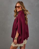 Woman in a burgundy knit turtleneck poncho sweater with lace-trim mini skirt, tinted aviator sunglasses and gold bangles