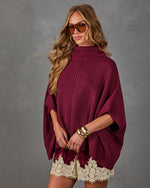 Woman wearing a burgundy turtleneck poncho sweater with gold bangles, tinted aviator sunglasses and cream lace-trim shorts