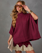 Woman wearing a burgundy knit turtleneck poncho sweater with velvet lace-trim shorts, gold cuff bracelets and tinted sunglasses
