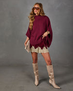 Woman in burgundy turtleneck poncho sweater, velvet lace-trim shorts, and tan knee-high suede boots holding a beige clutch