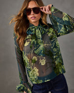 Woman wearing a sheer green floral chiffon blouse with neck bow and ruffle cuffs, aviator sunglasses and dark flare jeans