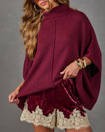 Burgundy knit turtleneck poncho sweater styled with crushed velvet shorts with cream lace trim and gold cuff bracelets