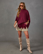 Woman in burgundy turtleneck poncho sweater, crushed velvet shorts with cream lace trim, and tan knee-high suede boots