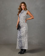A woman models a sleeveless, high-neck maxi dress with a black and white abstract spotted pattern, paired with black ankle boots, against a textured gray studio backdrop.