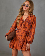A woman modeling a rust-orange long-sleeve mini dress with a dark floral print, holding a brown woven clutch against a neutral gray studio background.