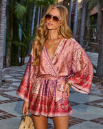 Woman wearing pink floral boho wrap mini dress with bell sleeves and tie waist, styled with aviator sunglasses and straw bag