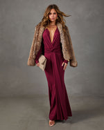 Woman in a burgundy long-sleeve plunging maxi dress with twist waist, styled with a faux fur coat and woven beige clutch