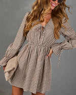 A woman wearing a brown and beige gingham mini dress with long sleeves, a tie neckline, and a gathered waist. She is holding a matching textured clutch against a plain gray studio background.