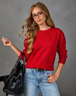 A woman wearing a bright red crewneck sweater, blue jeans, and glasses, holding a black woven handbag against a textured gray studio background.