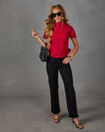 Woman in red pleated mock neck top with ruffle hem, black wide-leg jeans, strappy heels, sunglasses and black woven handbag