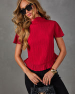 Woman wearing a red textured mock neck top with flutter sleeves and ruffle hem, black pants, sunglasses, and a woven black handbag