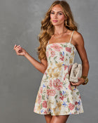 Woman wearing ivory floral mini dress with spaghetti straps, pink roses print, holding woven clutch, gold jewelry