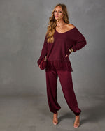 Woman in burgundy lace-hem V-neck sweater, satin jogger pants, velvet clutch and clear heels against a gray studio backdrop