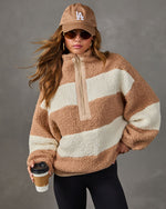 Woman wearing a tan and cream striped sherpa half-zip pullover with black leggings, an LA cap, sunglasses, and coffee cup