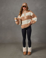 Woman in tan and cream striped sherpa quarter-zip pullover, black leggings, fuzzy platform boots, LA cap and sunglasses holding coffee
