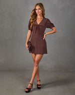 A woman models a short-sleeve brown mini dress with a white micro-dot pattern, a V-neckline, and a matching fabric tie at the chest. She is wearing black platform sandals and holding a small brown clutch, standing against a textured gray studio backdrop.