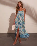 Woman wearing blue paisley floral halter maxi dress with front slit, clear heels, and white woven bag