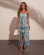 Woman wearing blue floral pleated halter maxi dress with front slit, styled with nude heels and white woven handbag