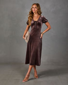 Woman in a brown velvet midi dress with lace neckline, satin bow and puff sleeves, styled with clear heels and a woven clutch