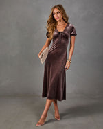 Woman wearing a brown velvet midi dress with lace trim, bow detail and puff sleeves, styled with clear heels and a woven clutch