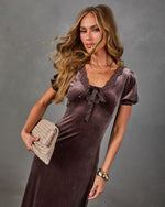 Woman in a brown velvet midi dress with lace trim, satin bow and puff sleeves, holding a woven beige clutch