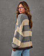 Woman in oversized gray and cream striped knit sweater with braided trim, worn with light wash jeans and aviator glasses