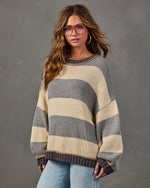 Woman wearing an oversized gray and cream striped knit sweater with braided trim, light wash jeans, and tinted glasses