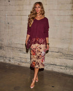 Woman in burgundy V-neck sweater and floral lace-trim satin midi skirt with clear heels, standing against a concrete wall