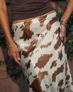 Woman wearing brown and white cow print satin midi skirt with dark brown crop top, silver bangles, outdoor setting