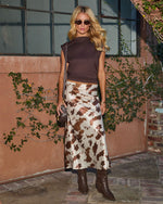 Blonde woman in brown crop top, cow print satin midi skirt, dark cowboy boots, and sunglasses posing outdoors