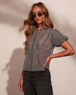 Woman wearing black gingham check short-sleeve button-up blouse, dark jeans, gold sunglasses, long wavy blonde hair