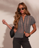Woman wearing gingham check sheer blouse, dark jeans, gold sunglasses, hoop earrings, holding black woven leather bag