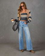 A woman stands against a gray studio backdrop wearing a beige and gray patterned knit cardigan over a matching ribbed tube top, light blue wide-leg jeans, and tan ankle boots. She holds a black woven tote bag and a takeaway coffee cup.