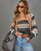 A woman models a cozy knit outfit featuring a beige, gray, and cream patterned cardigan, a matching strapless tube top, and light-wash blue jeans. She is accessorized with sunglasses, a woven gray shoulder bag, and holds a coffee cup against a neutral gray studio background.
